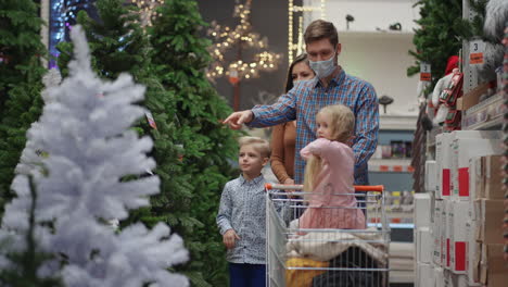 The-family-chooses-a-Christmas-tree-in-the-store-together.-A-happy-family-in-medical-masks-in-the-store-buys-Christmas-decorations-and-gifts-in-slow-motion